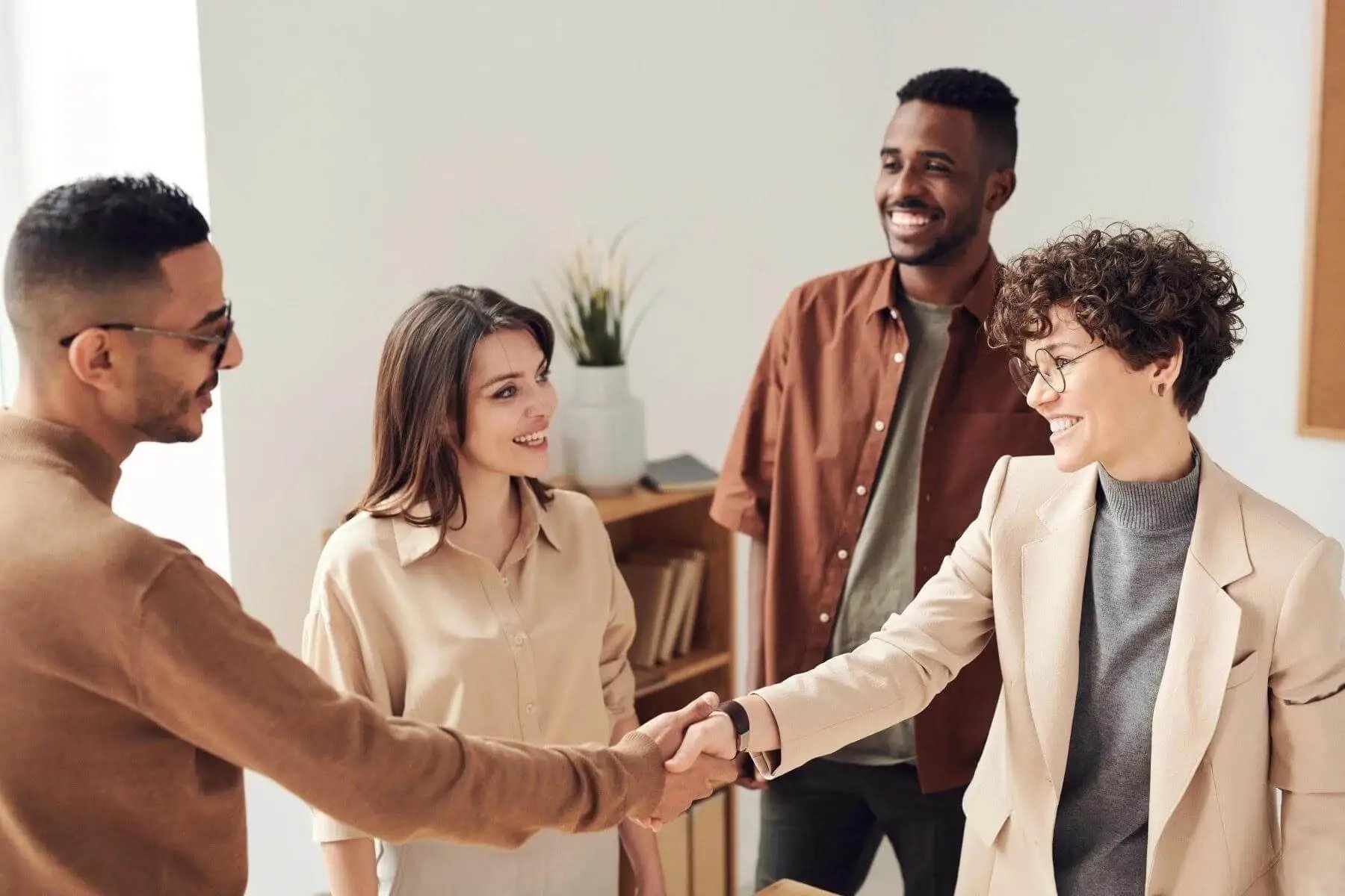 A diverse group of business people are greeting each other. Two of them, a man and a woman, are shaking hands. The other two, also a man and a woman, are smiling in greeting