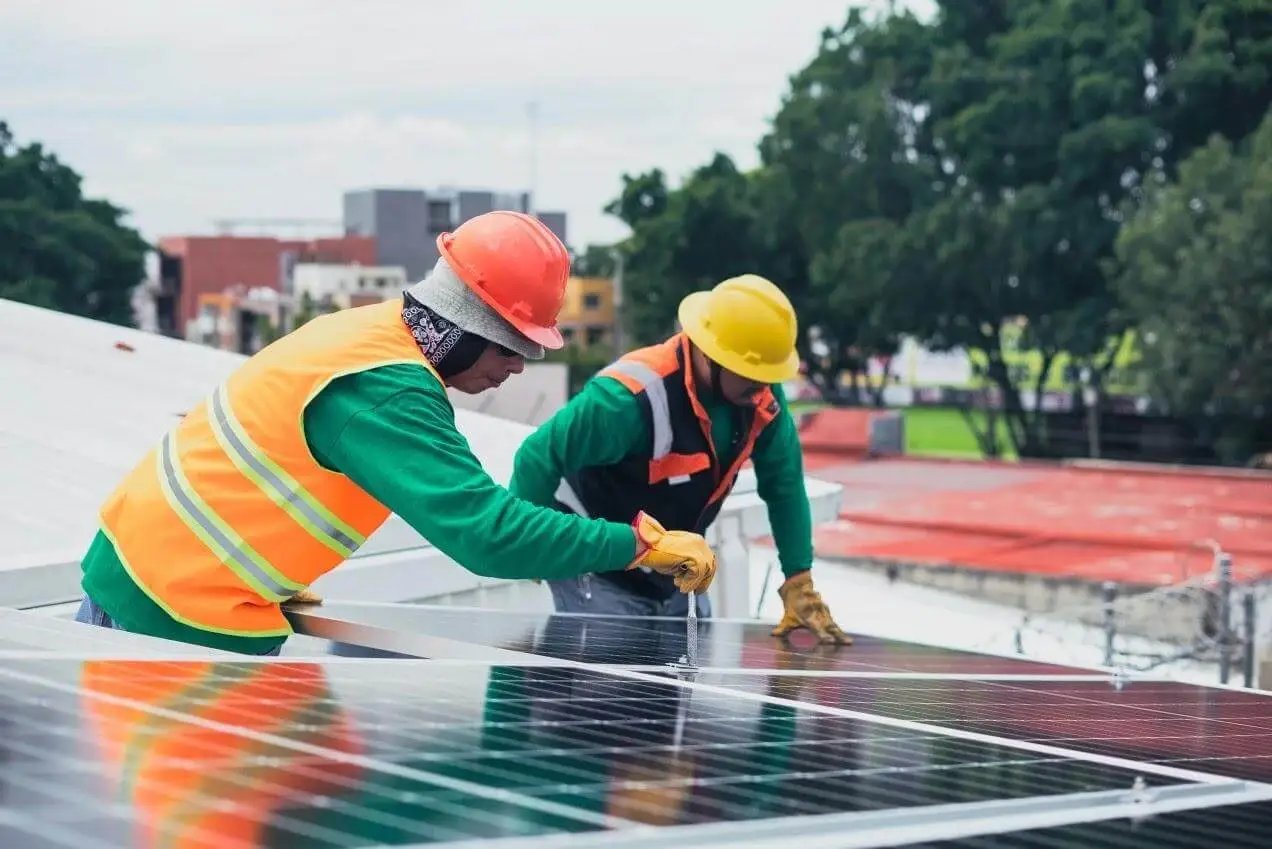 Two workpeople wearing hard hats and high visibility clothing install solar panels